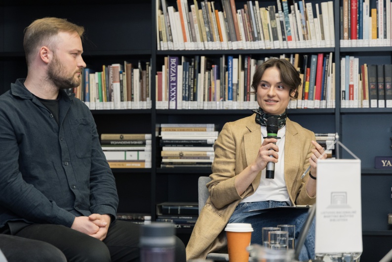 Diskusijos dalyviai, fone matosi knygų lentyna